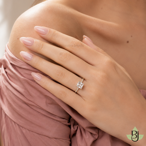 A realistic close-up photograph of a woman's fair-skinned hand resting gently on her bare shoulder, fingers relaxed and slightly overlapping with the thumb tucked underneath, displaying neatly manicured almond-shaped nails coated in a glossy light blush-pink polish that catches soft highlights, wearing a uploaded ring on the ring finger against a softly gathered dusty-rose pink fabric garment with gentle ruching and natural folds draping across the upper chest and shoulder area, smooth skin texture visible with subtle natural pores and warmth, soft diffused natural window light entering from the upper left creating gentle highlights on the nail surfaces and diamond facets while casting delicate shadows beneath the fingers and along the fabric creases, shot on a Canon EOS R5 with an 85mm f/1.8 lens at close focusing distance, shallow depth of field producing a smooth creamy bokeh on the out-of-focus background skin and fabric, natural color grading with warm soft tones and gentle contrast, high-resolution detail on the metallic ring sheen, diamond sparkle, and fabric texture, professional jewelry and beauty photography style, clean composition with the ring as the clear focal point. output image quality 2000 x 2000 pixel.